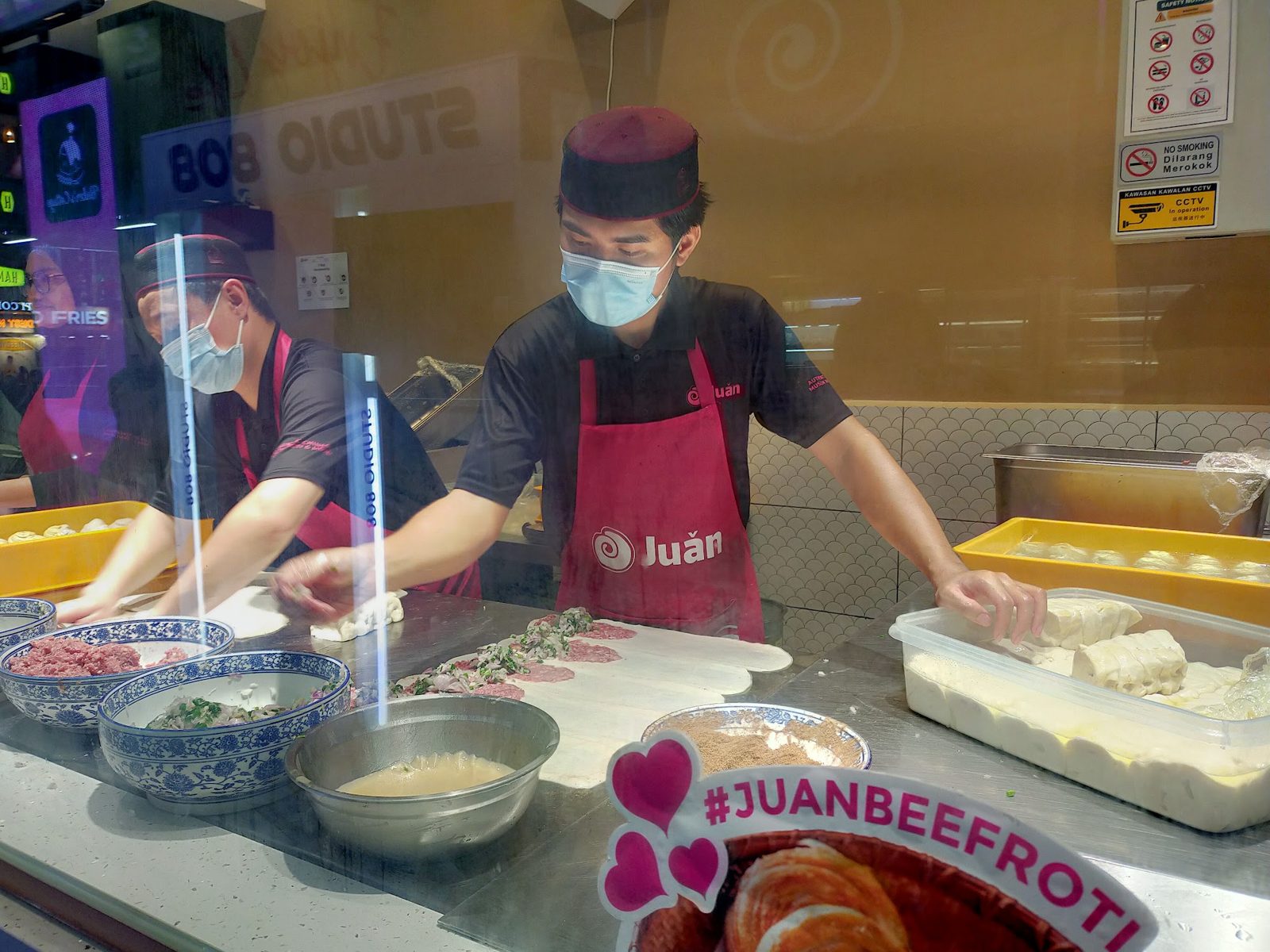 Juǎn crew in magenta aprons rolling roti behind the kiosk glass