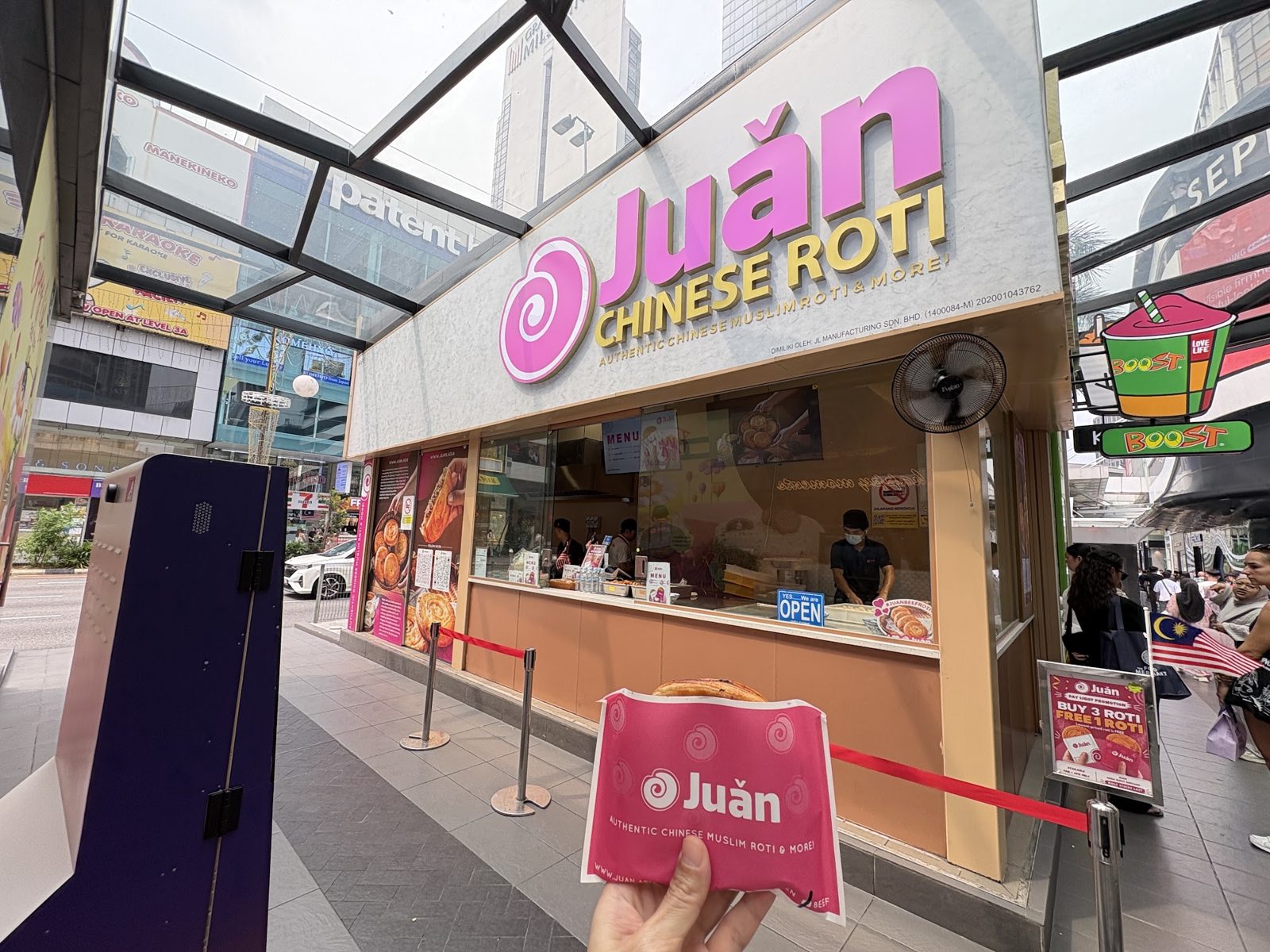 The Juǎn kiosk by day with its queue rope and a roti held in the foreground