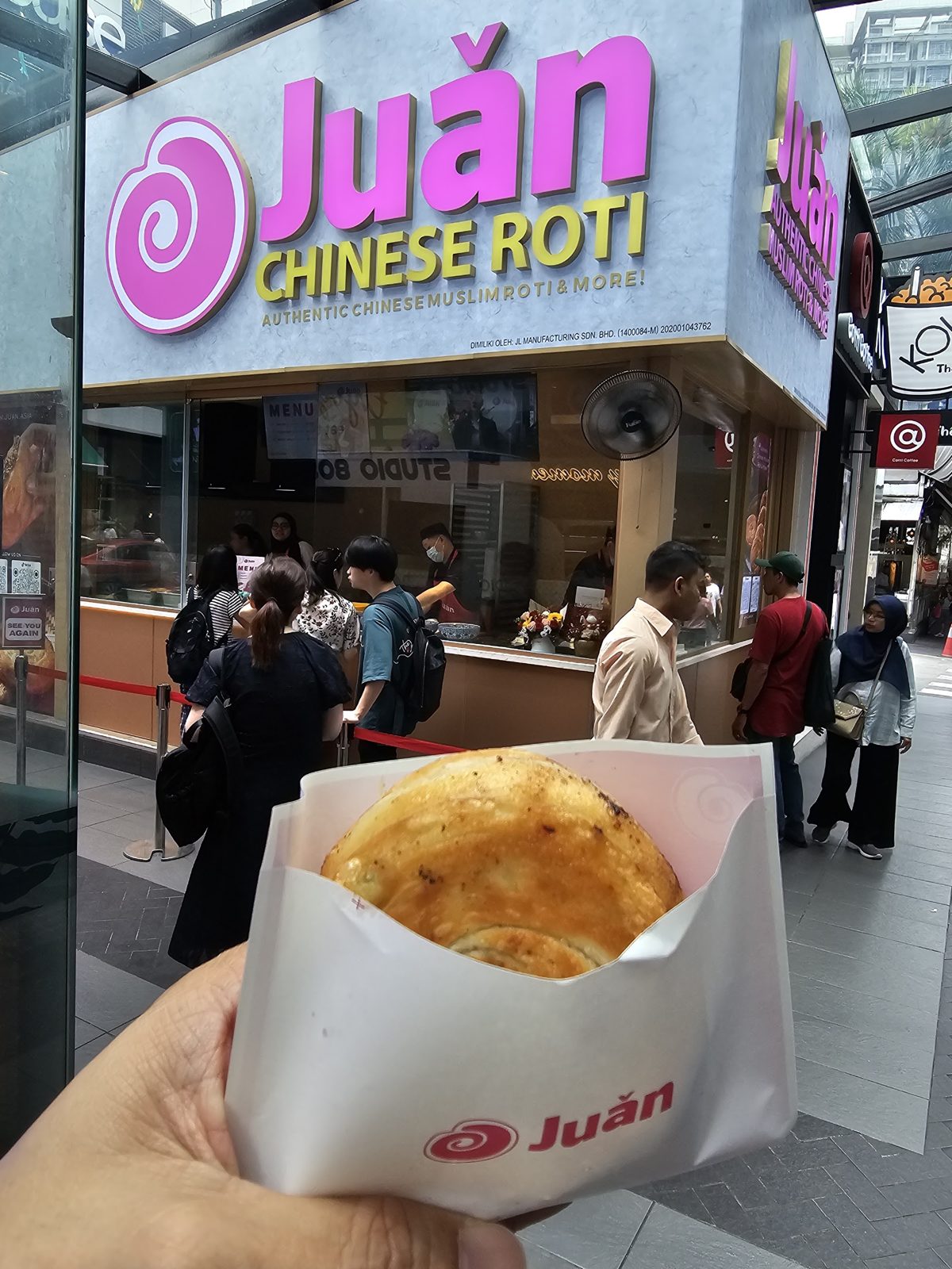 A golden Juǎn roti in its branded paper sleeve, held up in front of the kiosk